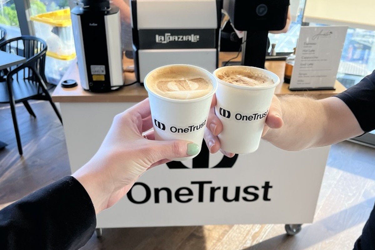 Two people raise OneTrust-branded coffee cups in front of a café espresso machine.