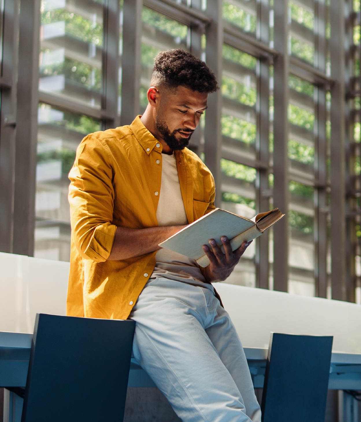 Un jeune homme dans des locaux professionnels qui lit un livre dans un hall de bureau éclairé par la lumière naturelle.